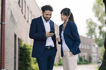 Businessman using smart phone standing by female colleague standing in office park - JOSEF02672