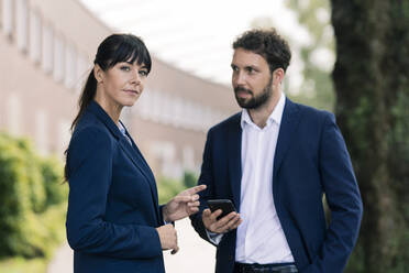 Confident businesswoman standing by male colleague holding smart phone - JOSEF02674