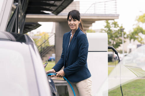 Lächelnde Unternehmerin beim Einstecken des Ladegeräts in ein Elektroauto - JOSEF02683