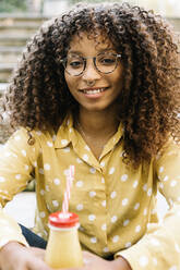 Curly hair woman with juice smiling while sitting outdoors - XLGF00792