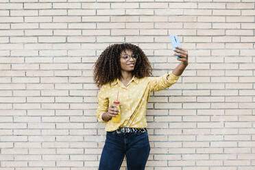 Junge Frau lächelnd bei der Aufnahme von Selfie durch Handy stehen gegen Backsteinmauer - XLGF00801