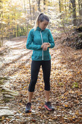 Sportswoman checking time while standing on forest path - JSRF01242