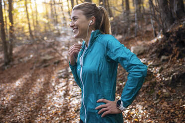 Smiling athlete listening music while standing with hand on hip in forest - JSRF01253