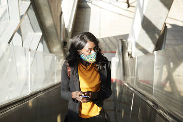 Thoughtful female entrepreneur with mobile phone on escalator during COVID-19 - PMF01615