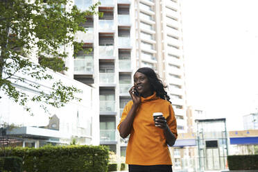 Smiling businesswoman with disposable cup talking on phone at office park - PMF01642