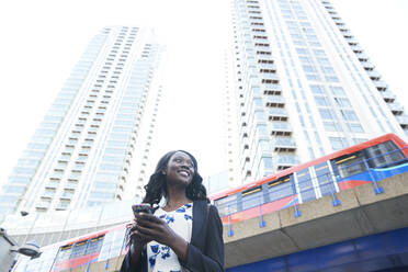 Smiling businesswoman with mobile phone against metro train and skyscrapers - PMF01653