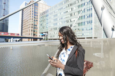 Lächelnde Geschäftsfrau auf einer Brücke, die an einem sonnigen Tag ein Mobiltelefon benutzt - PMF01657