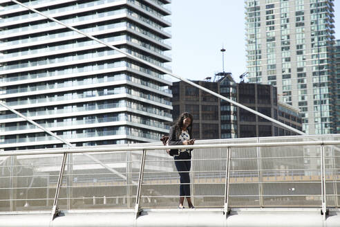 Geschäftsfrau, die ein Mobiltelefon benutzt, während sie auf einer Brücke in der Stadt steht - PMF01662
