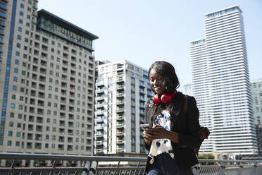 Geschäftsfrau mit Rucksack und Kopfhörern, die ein Mobiltelefon benutzt, während sie auf einer Brücke in der Stadt spazieren geht - PMF01670