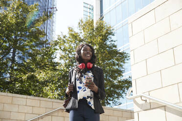 Smiling businesswoman with headphones and mobile phone carrying backpack while walking in city - PMF01676