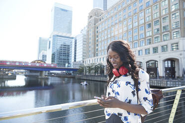 Businesswoman with backpack and headphones using mobile phone while standing on bridge in city - PMF01682