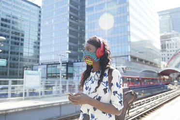 Businesswoman wearing face mask and headphones looking away while standing at station - PMF01689