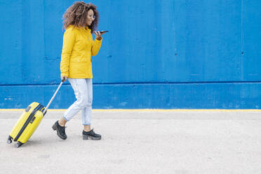 Frau mit Gepäck, die mit einem Mobiltelefon spricht, während sie gegen eine blaue Wand läuft - JCZF00347