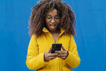 Überraschte Frau mit offenem Mund, die ein Mobiltelefon benutzt, während sie an einer blauen Wand steht - JCZF00349