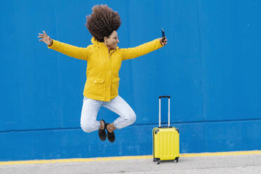 Young carefree woman jumping while taking selfie through mobile phone against blue wall - JCZF00351