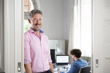 Lächelnder Geschäftsmann mit einem männlichen Kollegen im Hintergrund in einer Bürokabine - FKF03868