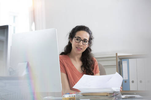 Weibliche Fachkraft, die ein Papierdokument hält und auf einen Computer im Büro schaut - FKF03924