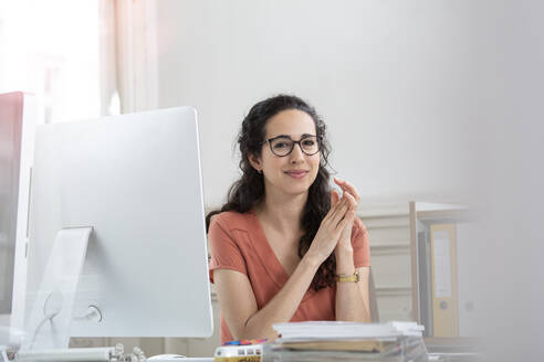 Female professional with hands clasped sitting by computer - FKF03926