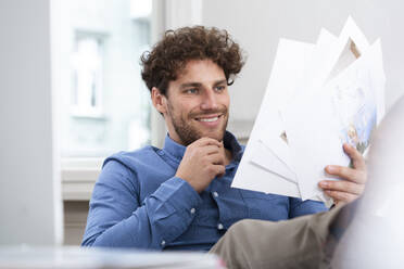 Smiling businessman with hand on chin reading document in office - FKF03932