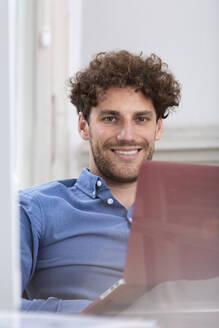 Männlicher Berufstätiger, der lächelnd mit einem Laptop in einer Bürokabine sitzt - FKF03937