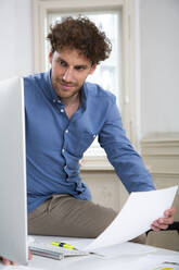 Young businessman working on computer with document at desk in office - FKF03940