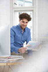 Männlicher Berufstätiger, der am Computer sitzend im Büro ein Dokument liest - FKF03941