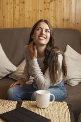 Nachdenkliche Frau lächelt, während sie zu Hause auf dem Sofa sitzt - LJF01871