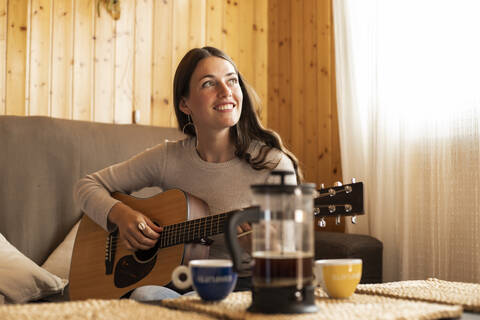 Thoughtful young woman playing guitar while sitting on sofa at home stock photo