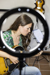 Junge Frau mit Webcam-Licht spielt Gitarre, während sie zu Hause sitzt - LJF01897