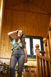 Schöne junge Frau steht mit der Hand im Haar an einer Treppe in einem Haus - LJF01905