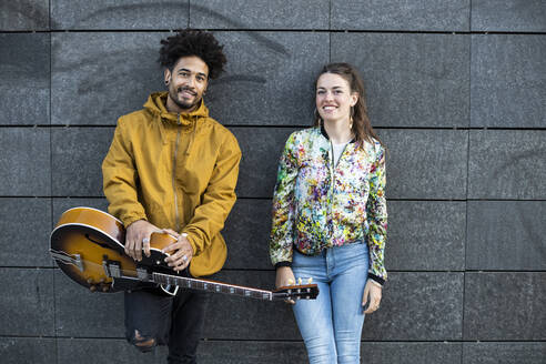Lächelnder Mann, der eine Gitarre hält, während er neben einer Frau an einer Betonwand steht - LJF01911