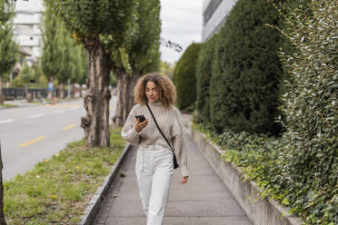 Junge Frau benutzt ihr Smartphone, während sie auf einem Fußweg in der Stadt spazieren geht - MEF00046