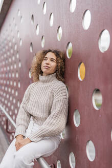 Nachdenkliche junge Afro-Frau sitzt auf einem Geländer vor einer kastanienbraunen Metallwand - MEF00054