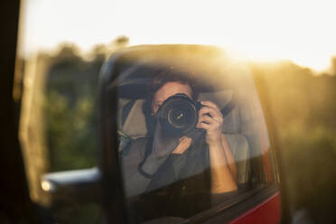 Side-view mirror reflection of woman photographing setting sun - MAMF01474