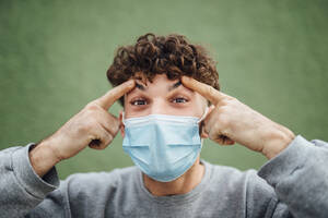 Young man touching eyebrows against green background in studio during COVID-19 - MIMFF00313