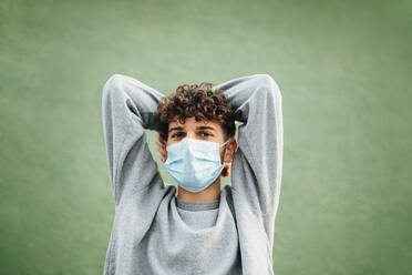 Caucasian man with hands behind head wearing surgical mask against green background - MIMFF00315