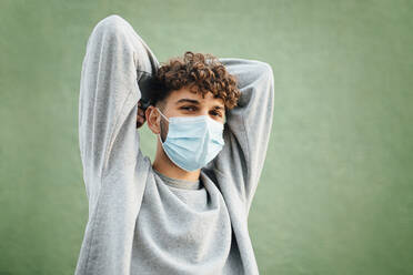 Young man with hands behind head against green background during coronavirus - MIMFF00316