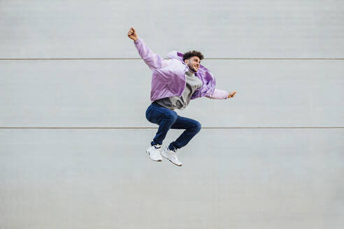 Carefree man with arms outstretched jumping against wall - MIMFF00320