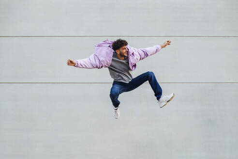 Young man jumping happily against wall - MIMFF00321