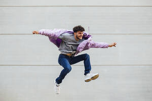 Carefree young man with arms outstretched jumping against gray wall - MIMFF00322