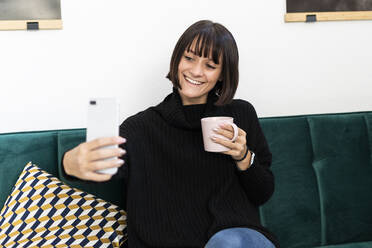 Lächelnde junge Frau macht Selfie mit Kaffeetasse im Wohnzimmer - GIOF09911