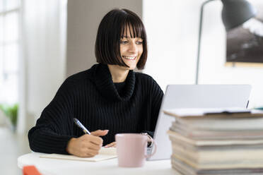 Smiling young female student with laptop writing while studying in study room - GIOF09927