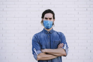 Junger Mann mit verschränkten Armen und Gesichtsschutzmaske vor einer weißen Backsteinmauer - EBBF01720