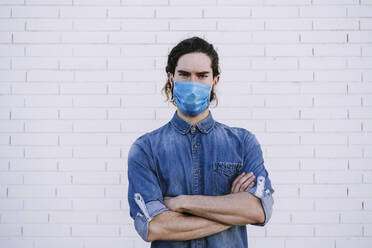 Young man with arms crossed wearing protective face mask against white brick wall - EBBF01720