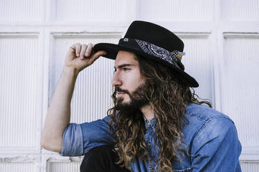 Stilvoller junger Mann mit cooler Einstellung, der einen Hut gegen eine weiße Wand hält - EBBF01722