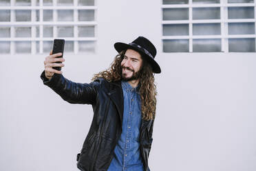 Glücklicher junger Mann in Lederjacke macht ein Selfie vor einer weißen Wand - EBBF01739