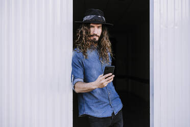 Confident young man with mobile phone leaning on corrugated wall in doorway - EBBF01743