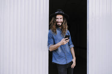 Happy stylish man leaning on corrugated wall while holding mobile phone in doorway - EBBF01744