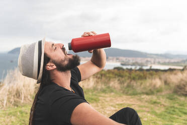 Junger Mann mit Hut trinkt mit geschlossenen Augen Wasser in Santander, Spanien - JAQF00015
