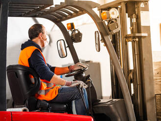 Side view unrecognizable male worker in high visibility vest and face mask driving autoloader machine while working in warehouse - ADSF18793